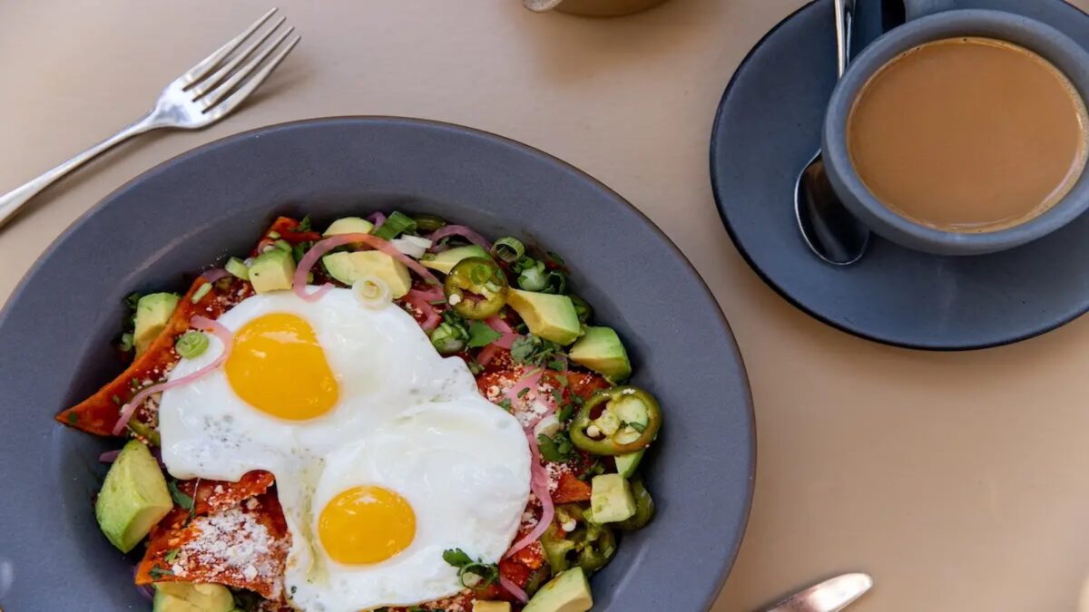 A colorful breakfast dish featuring two sunny-side-up eggs is served in a bowl with tomatoes, avocado, jalapeño slices, and green onions. A fork is placed beside the bowl, and there is a cup of coffee on a saucer next to the dish. Join the Clean Plate Club with this delightful morning meal!