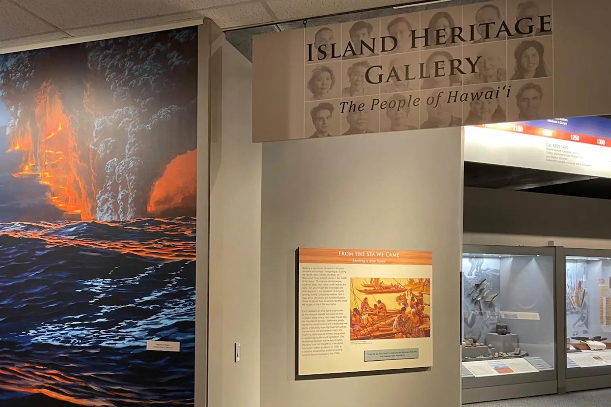 An image of the Island Heritage Gallery, one of the Best Places to Visit on the Big Island, featuring a large mural of a traditional Hawaiian canoe at sea near an erupting volcano. The entrance sign reads "Island Heritage Gallery: The People of Hawai'i." An informational display is visible below the sign.