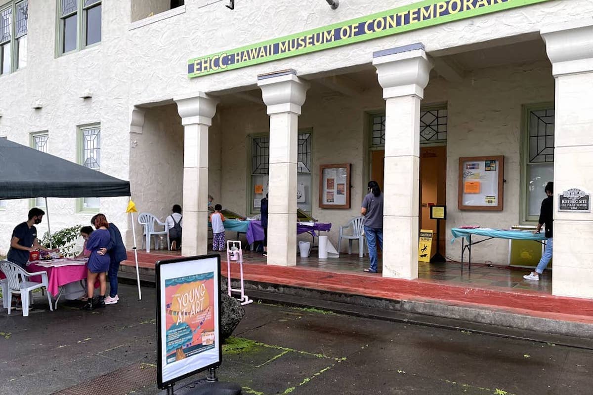 A building with white pillars and tables and signs set up with a sign above the entrance that reads "EHCC: Hawaii Museum of Contemporary."