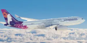 A Hawaiian Airlines plane with a colorful tail design featuring a woman's face and hibiscus flower is soaring above a blanket of clouds against a clear blue sky, with the word "HAWAIIAN" visible on the side. Passengers' carry on luggage is securely stored as they enjoy the stunning aerial view.