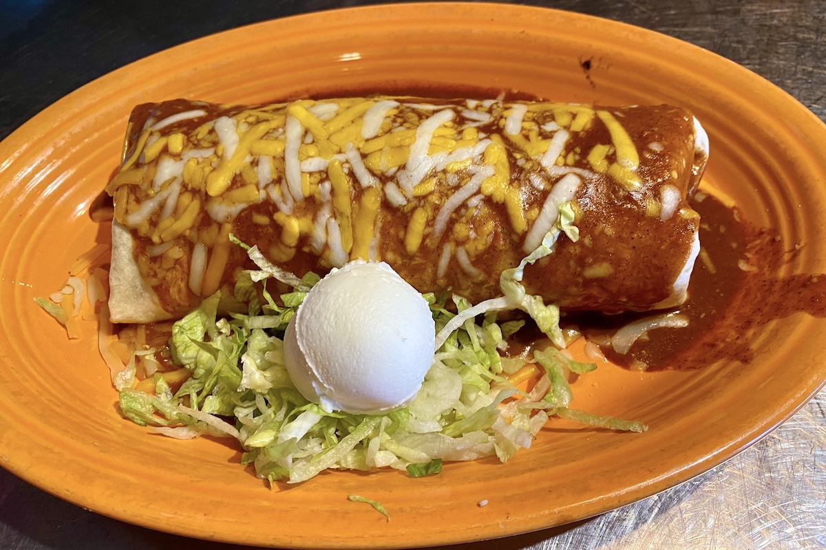 An enchilada topped with melted cheese and enchilada sauce sits on an orange plate. It is accompanied by shredded lettuce and a scoop of sour cream on the side, making it a contender for the Best Dinner in Maui.