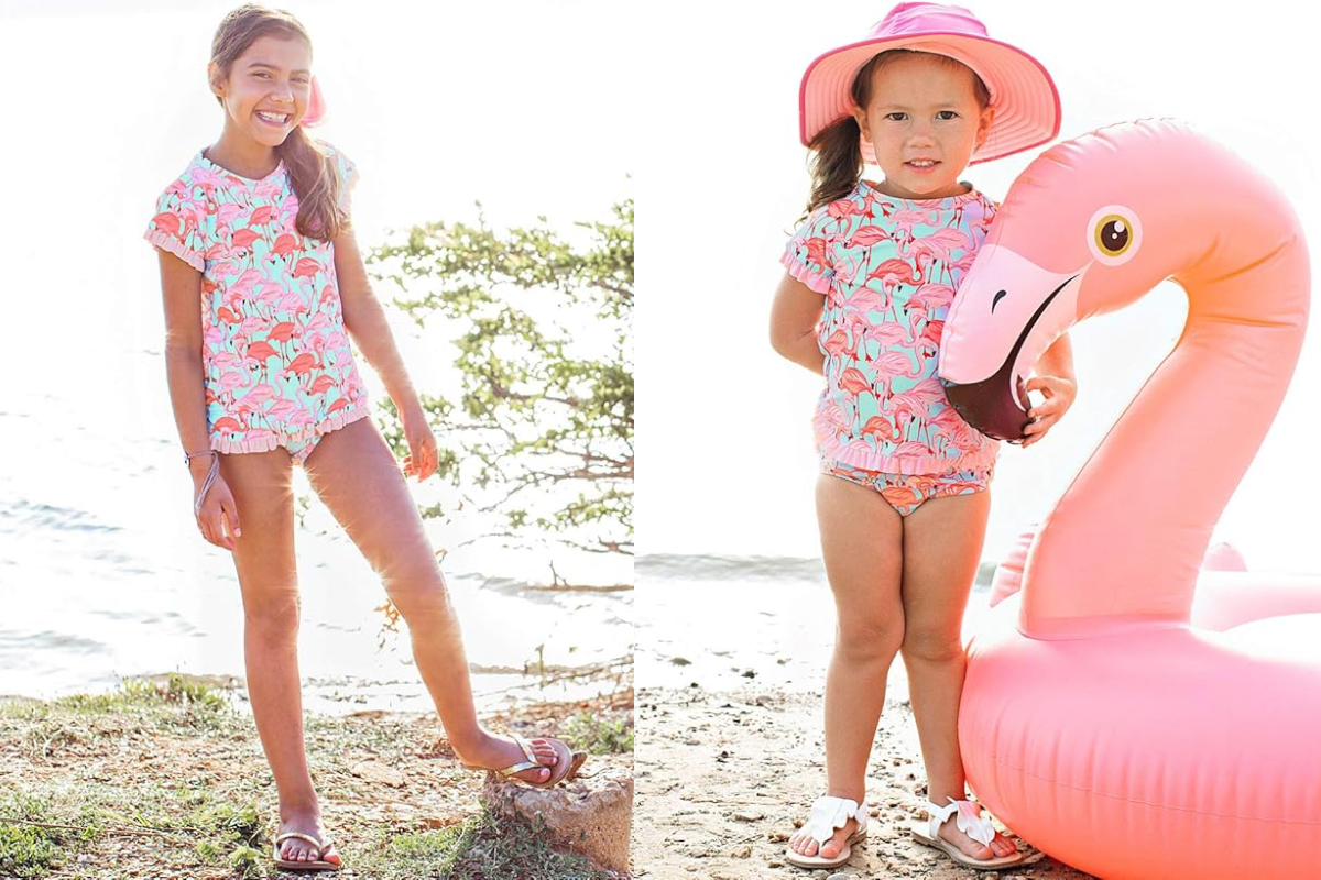 Two young children stand near a large inflatable pink flamingo float by the water. Both are wearing matching pink and blue flamingo-themed swimwear, complete with baby UV tops. The older child, on the left, is smiling, while the younger child, on the right, sports a pink hat.