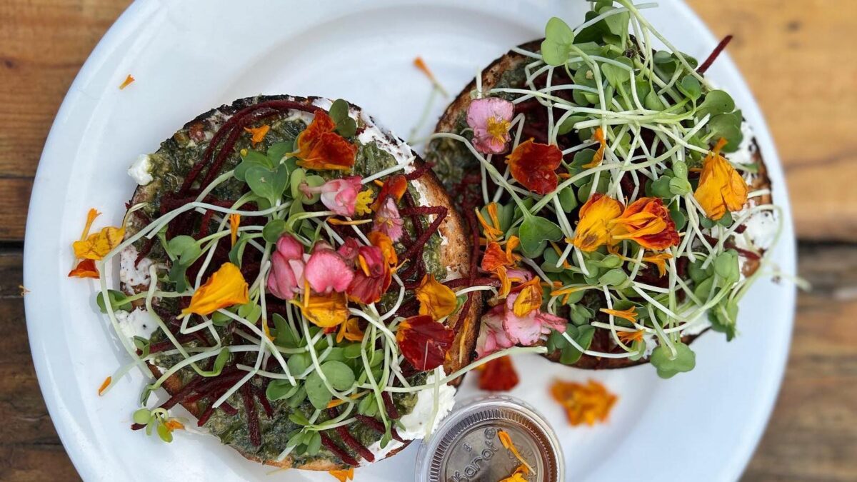 A white plate holds two colorful bagels topped with cream cheese, sprouts, microgreens, edible flowers, and shredded beets. A small container of dressing is placed beside the bagels. The vibrant ingredients create a visually appealing dish that's among the Best Breakfast Kauai offers.