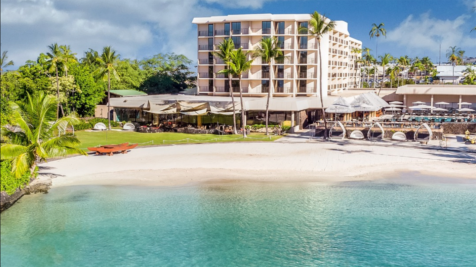 Image of a serene beachfront resort on the Big Island featuring a modern hotel building with multiple floors, lush green palm trees, outdoor dining areas, and various seating arrangements by the turquoise lagoon. White sand meets the clear blue water under a partly cloudy sky, epitomizing one of the best-vetted value hotels.