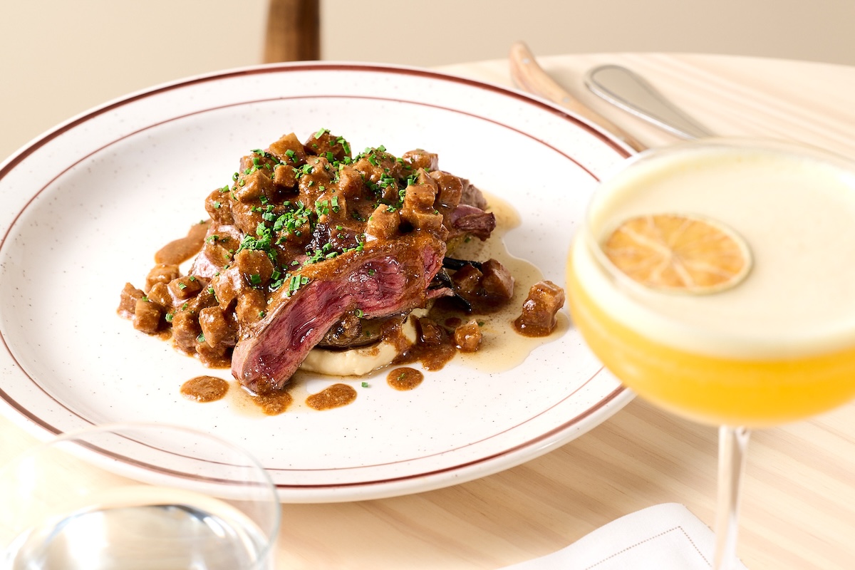 A plate with sliced beef in a sauce and topped with chopped herbs. A yellow cocktail is on the side.