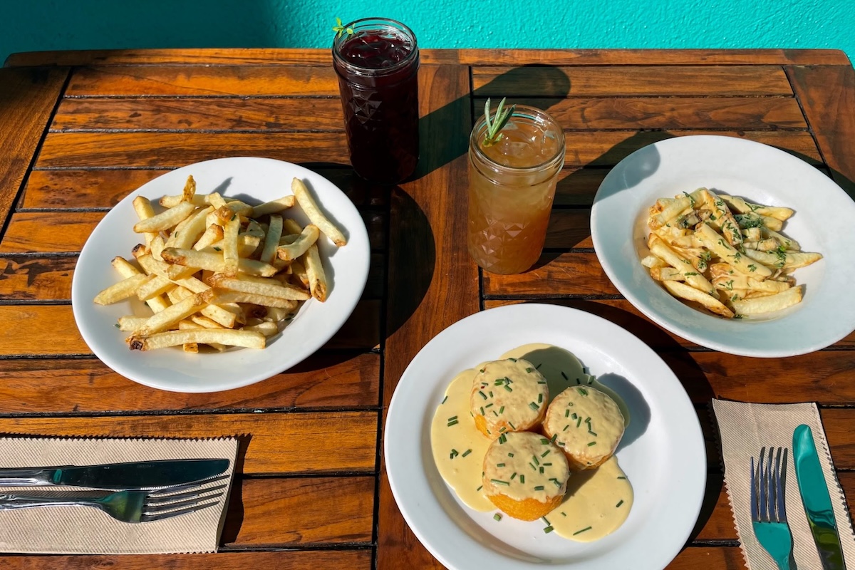 A wood table with a plate of fries, a plate of Eggs Benedict and a plate of cheesy fries, as well as two drinks.
