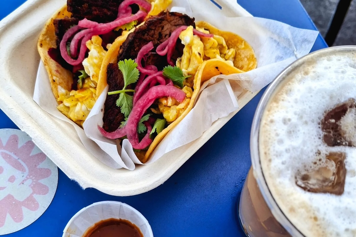 On a blue table sit tacos wrapped in paper and a coffee on the side.