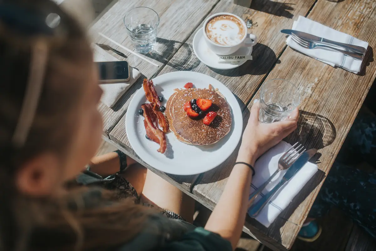 A person sits at a wooden table with a breakfast setting. They have a plate of pancakes topped with strawberries, accompanied by bacon, a glass of water, and a cup of coffee with foam art—the best breakfast North Bay has to offer. Their phone and a pair of sunglasses are also on the table.