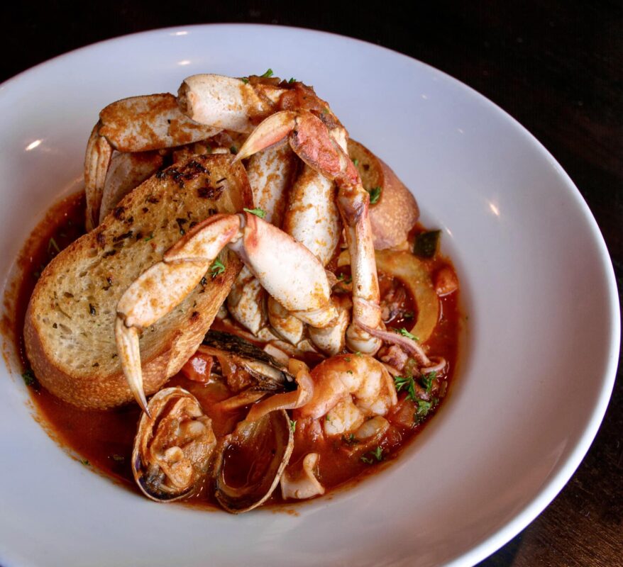 A white bowl filled with seafood cioppino, featuring crab legs, clams, shrimp, and mussels, is garnished with grilled bread slices. The dish is served in a rich tomato-based broth and sprinkled with fresh herbs—truly the best lunch South Bay has to offer.