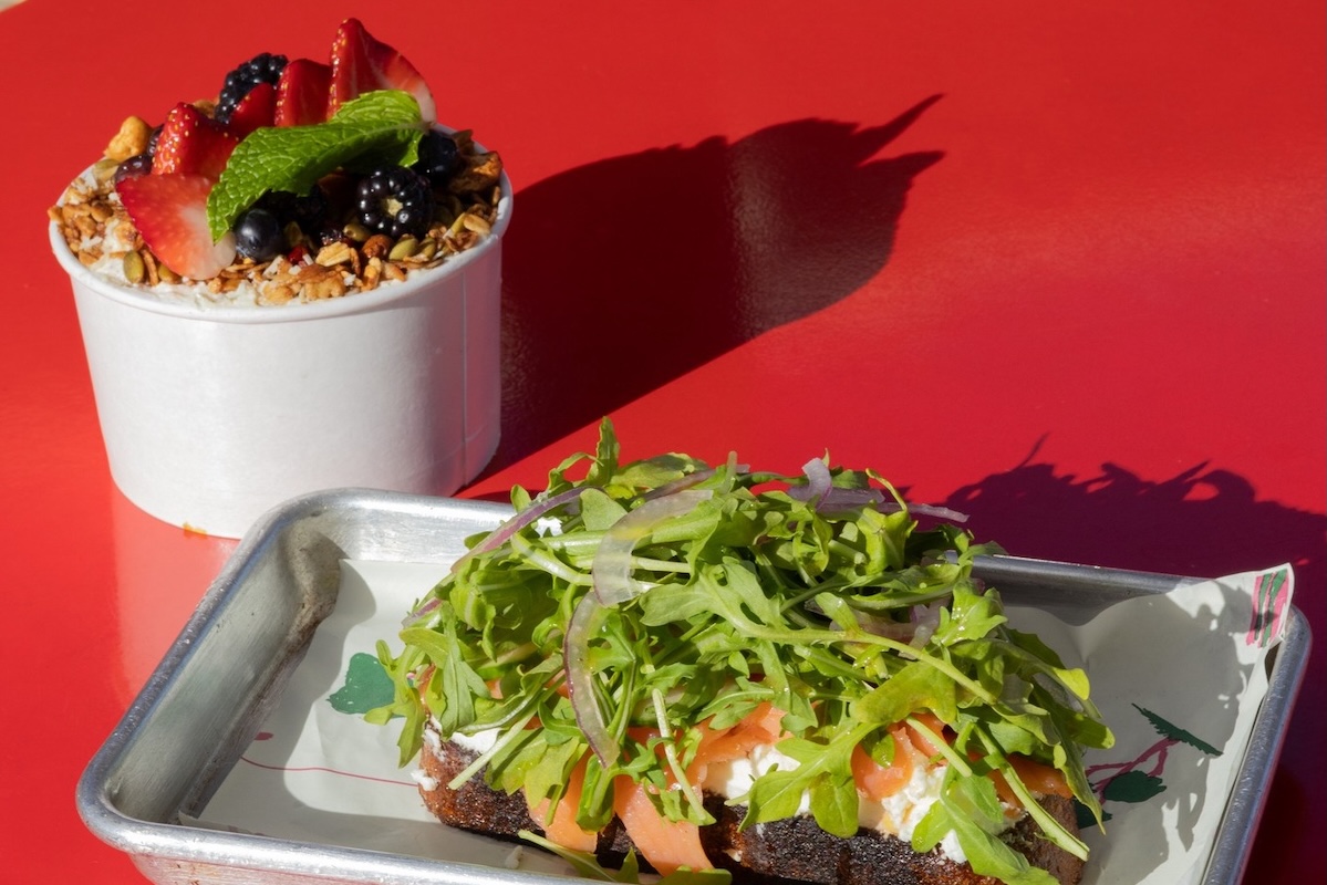 On a red table, a tray with toast topped with cheese, lox, arugula and red onion, and a parfait topped with granola, blackberries, blueberries, strawberries and mint for garnish.