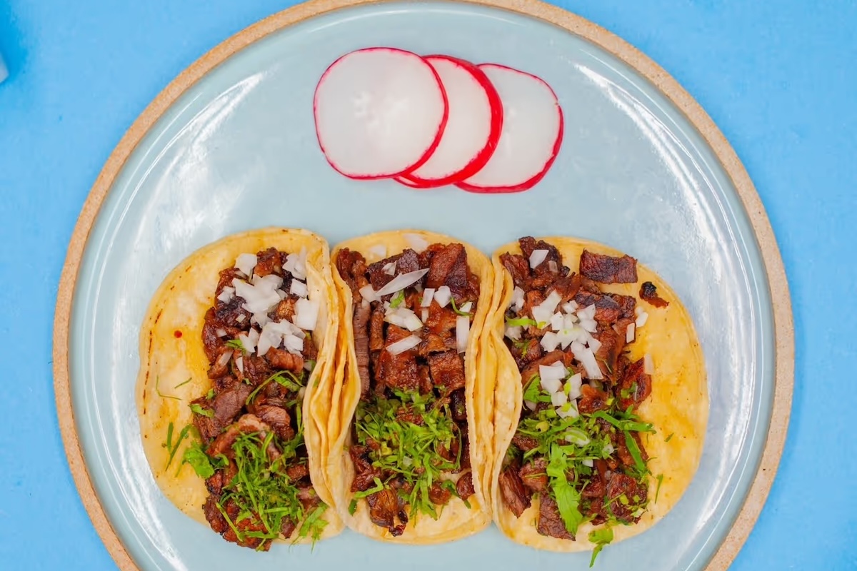 A blue background with a plate that has three tacos topped with chopped herbs and onions and slices of radish on the side.