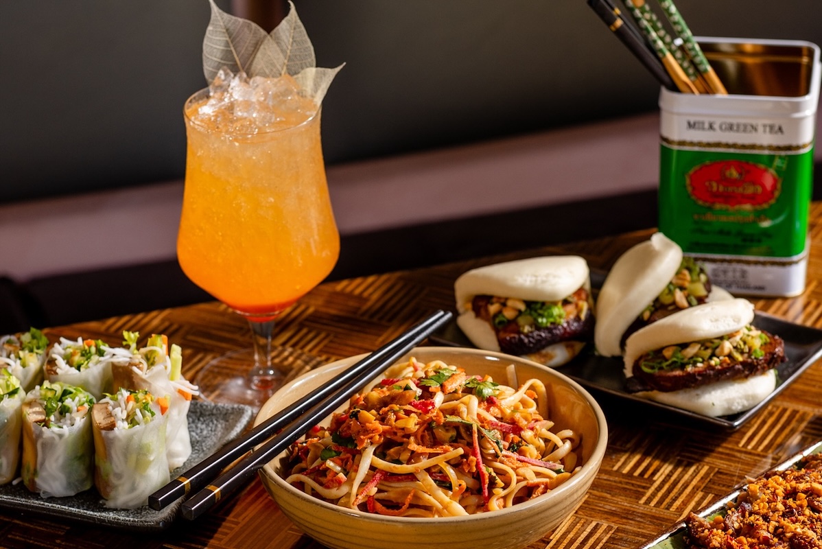 A table with a bowl of noodles centers, pork buns in the background, halves of spring rolls standing up to to the left, and an iced orange beverage with sheer leaves or garnish.