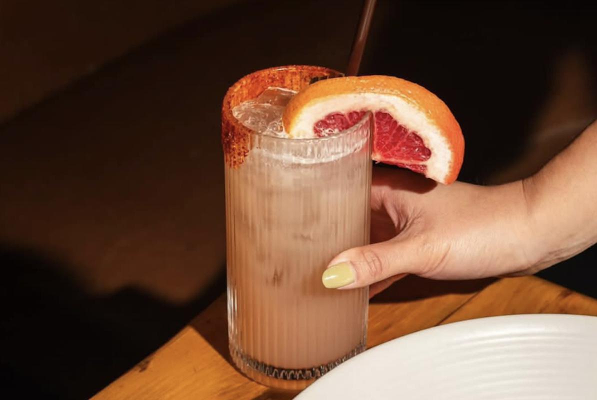 From off screen, awoman holds onto a cocktail sitting on table. It's a light pink color with a blood orange and reddish seasoning along the rim.