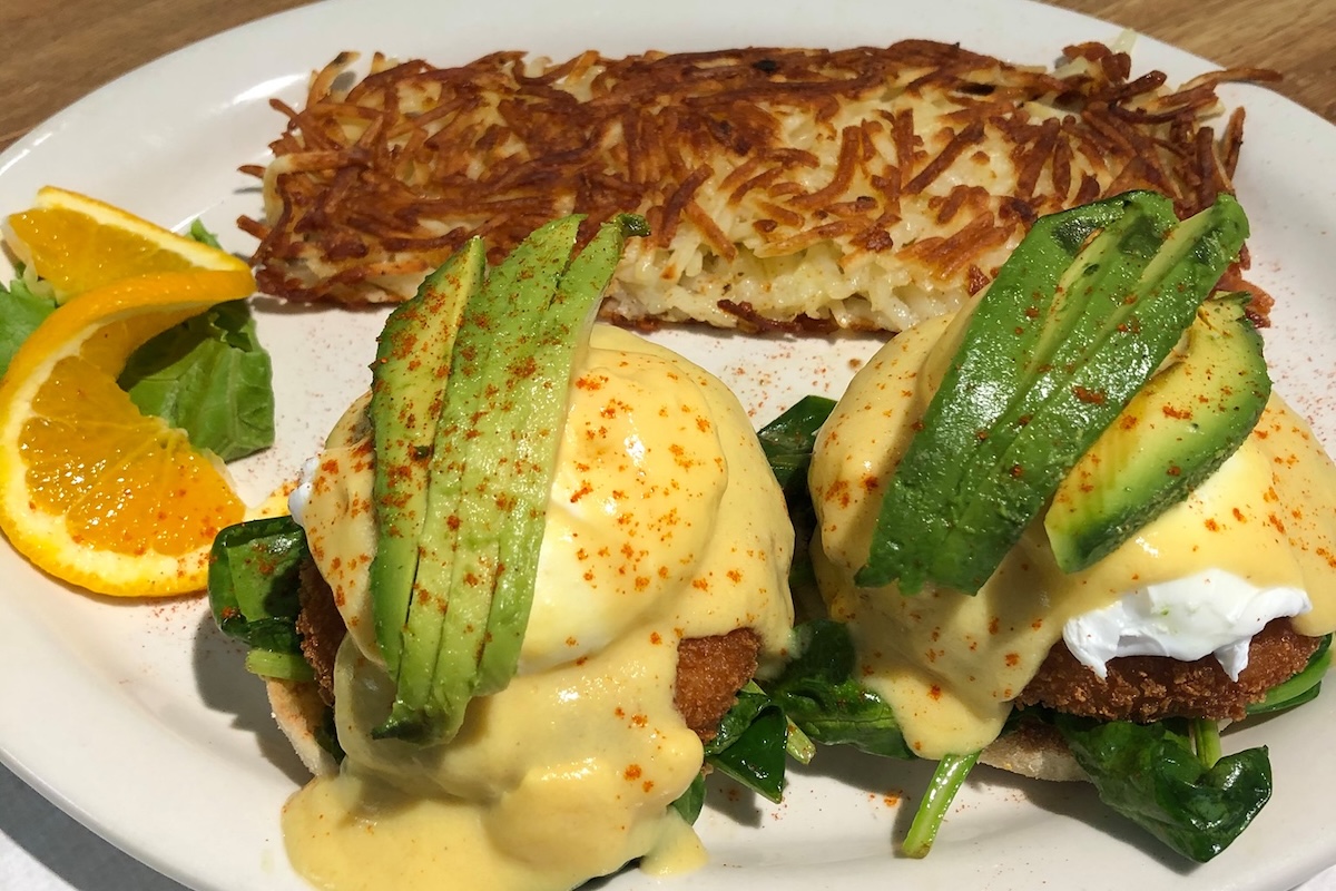 A plate with eggs benedict topped with avocado and a side of hash browns and orange slices.