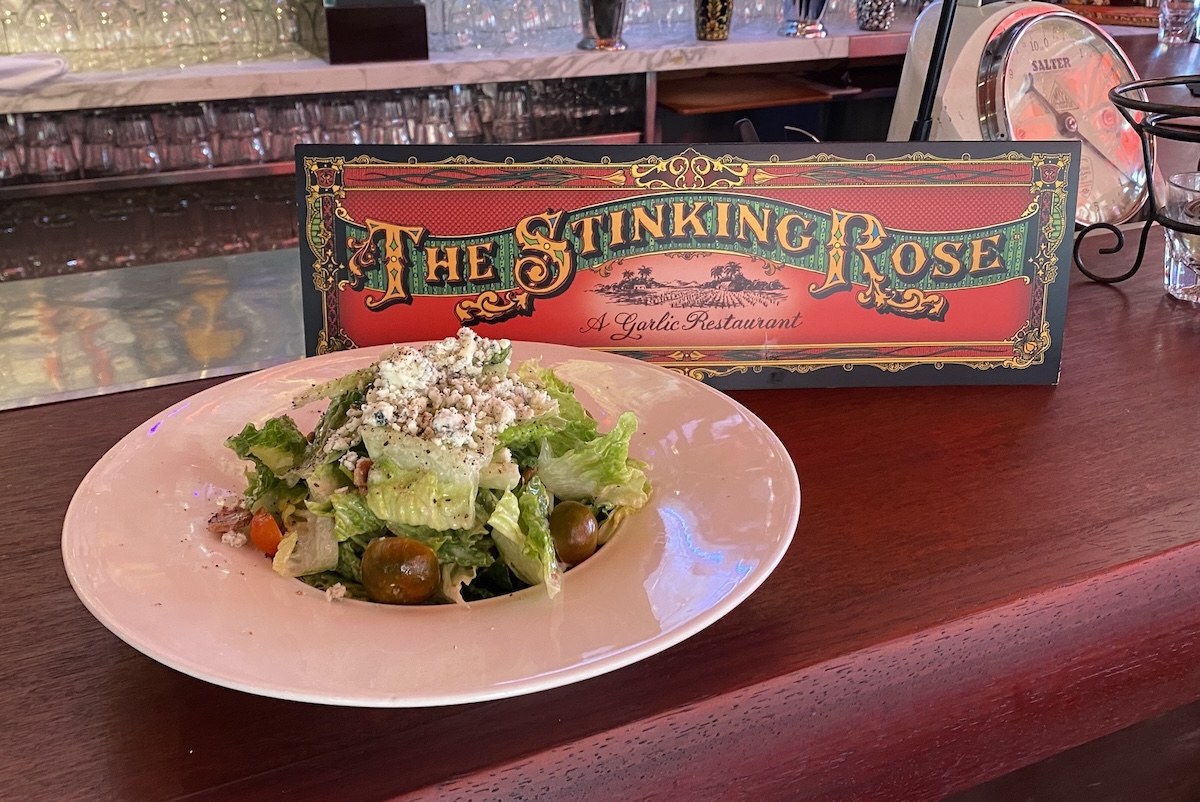 A plate of salad topped with blue cheese sits on a bar next to a sign that says "The Stinking Rose."