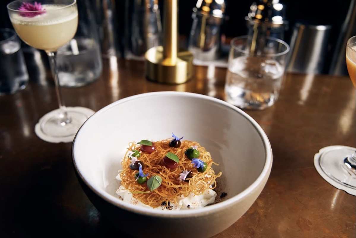 A white bowl with a bed of crunchy bit topped with dollops of sauce and leaves for garnish. A cocktail with a pink flower for garnish sits next to the bowl.