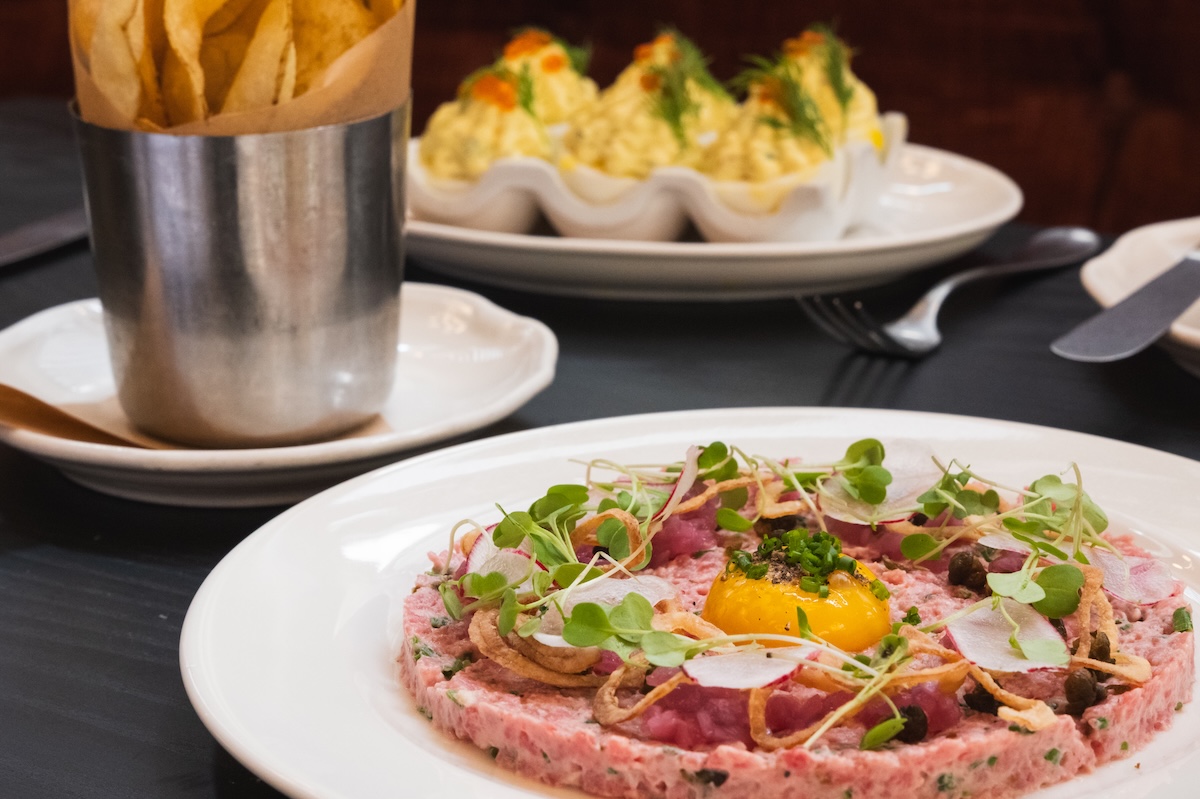 A plate with a flat pink disk of food topped with an egg yolk, crunchy pieces, microgreens and radish slices, and some hard boiled eggs and chips on the side.