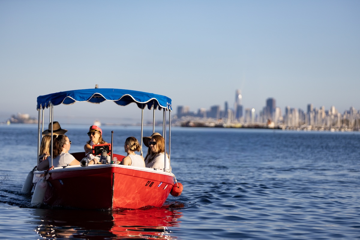 A red boat with a blue awning floats on the bay with a view of San Francisco in the background.