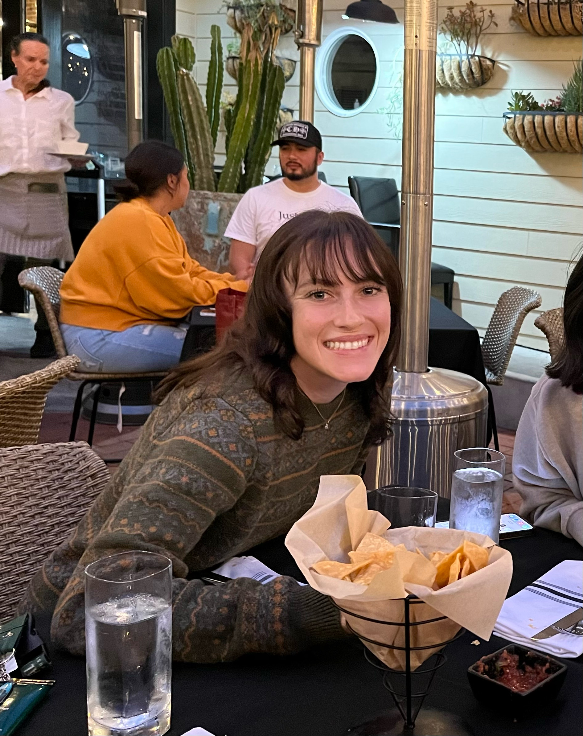 A person with long brown hair smiles while sitting at an outdoor restaurant table. The table has a basket of chips and glasses of water, perfect for any food editor exploring local getaways. Other diners are seated in the background, adding to the cozy setting adorned with plants and warm lighting.