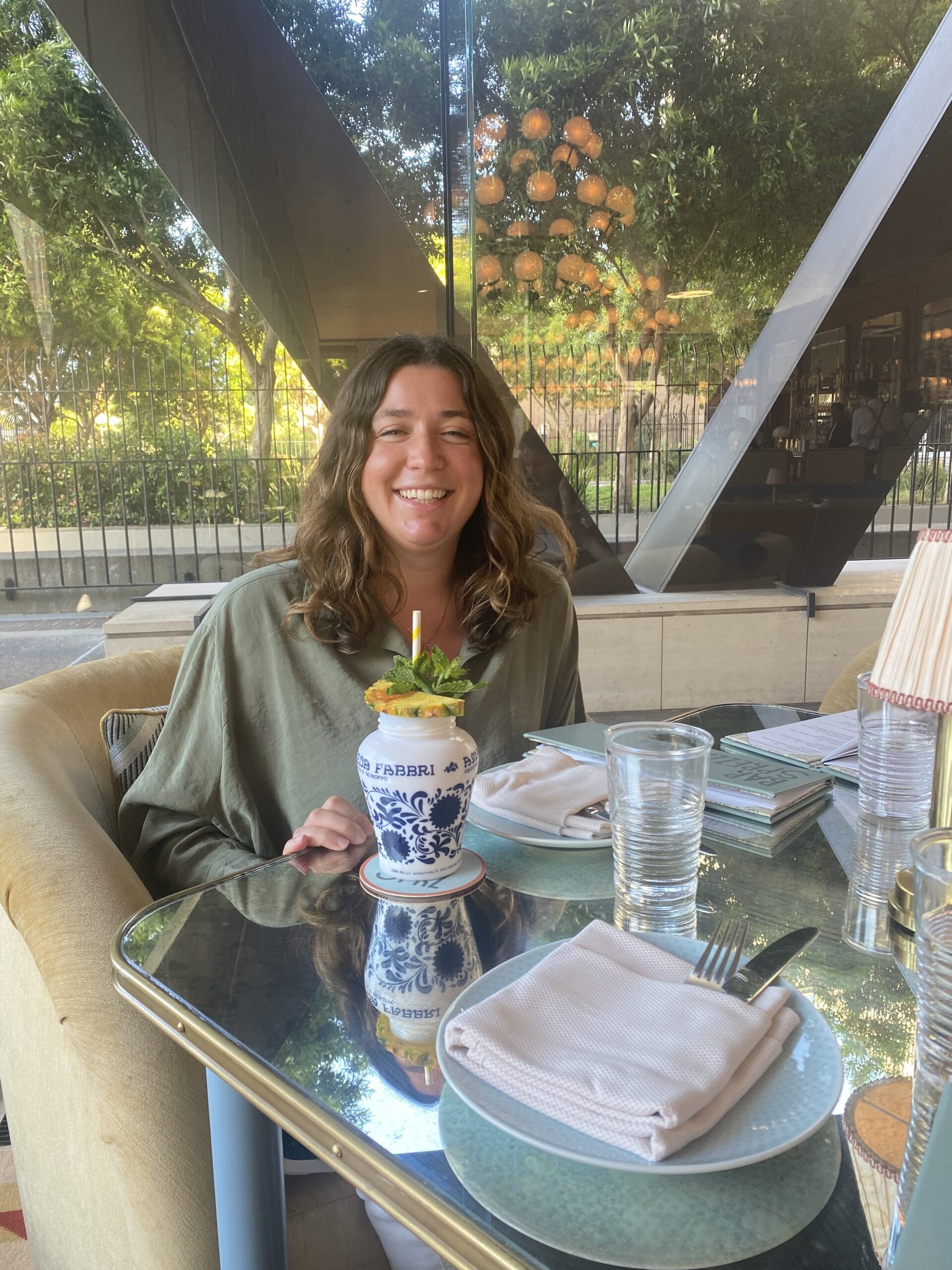 A person with long brown hair smiles at a table in a restaurant, recommended by local food editors. They are seated in front of a tropical dessert in a decorative cup, with plates, cutlery, and a glass of water on the table. The background shows large windows and green foliage outside.