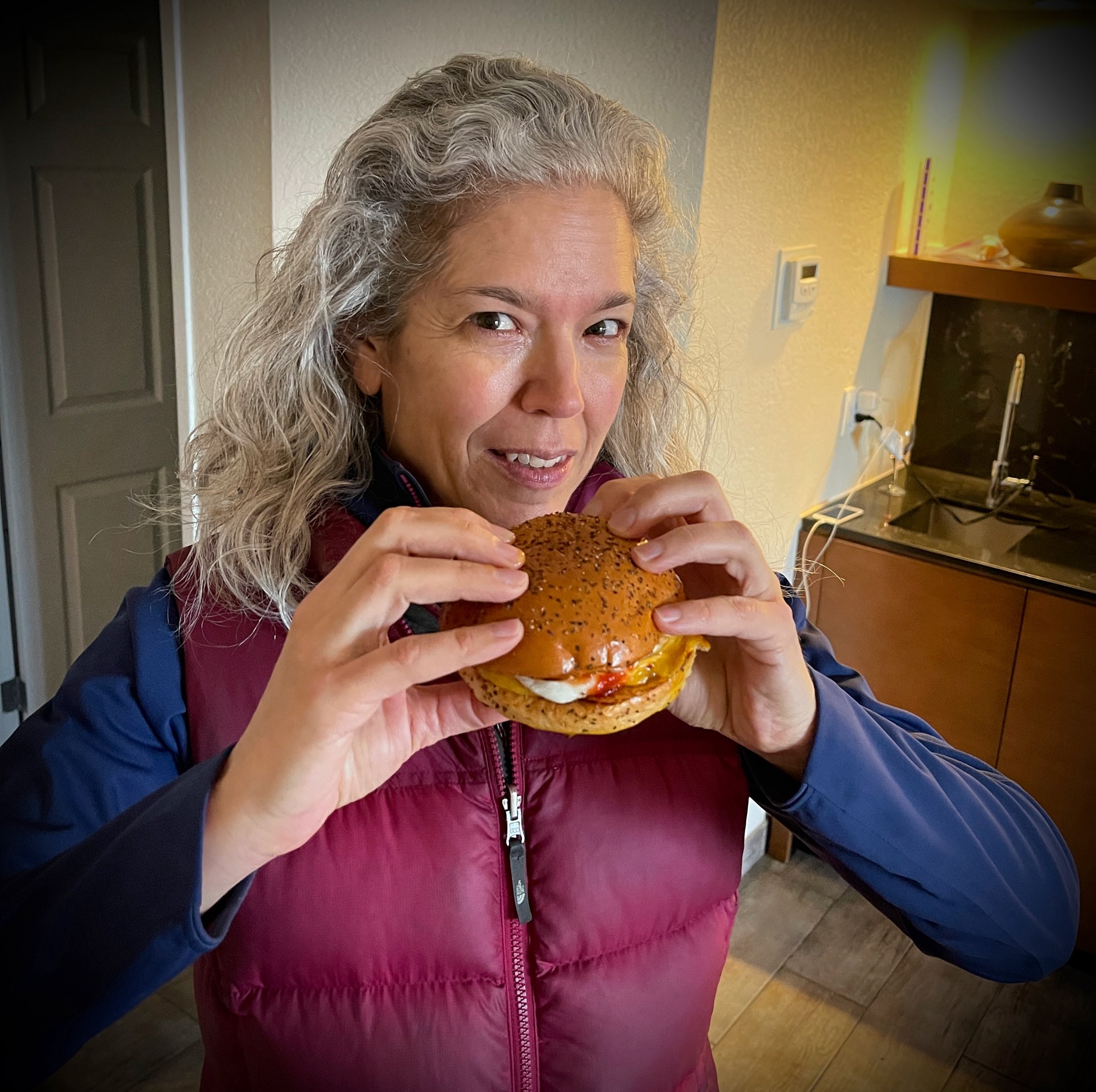 A person with long, wavy gray hair, wearing a maroon vest and a blue long-sleeve shirt, smiles and holds a sandwich with both hands in front of their face in a cozy indoor setting, reminiscent of photos seen in local getaways magazines curated by top food editors.