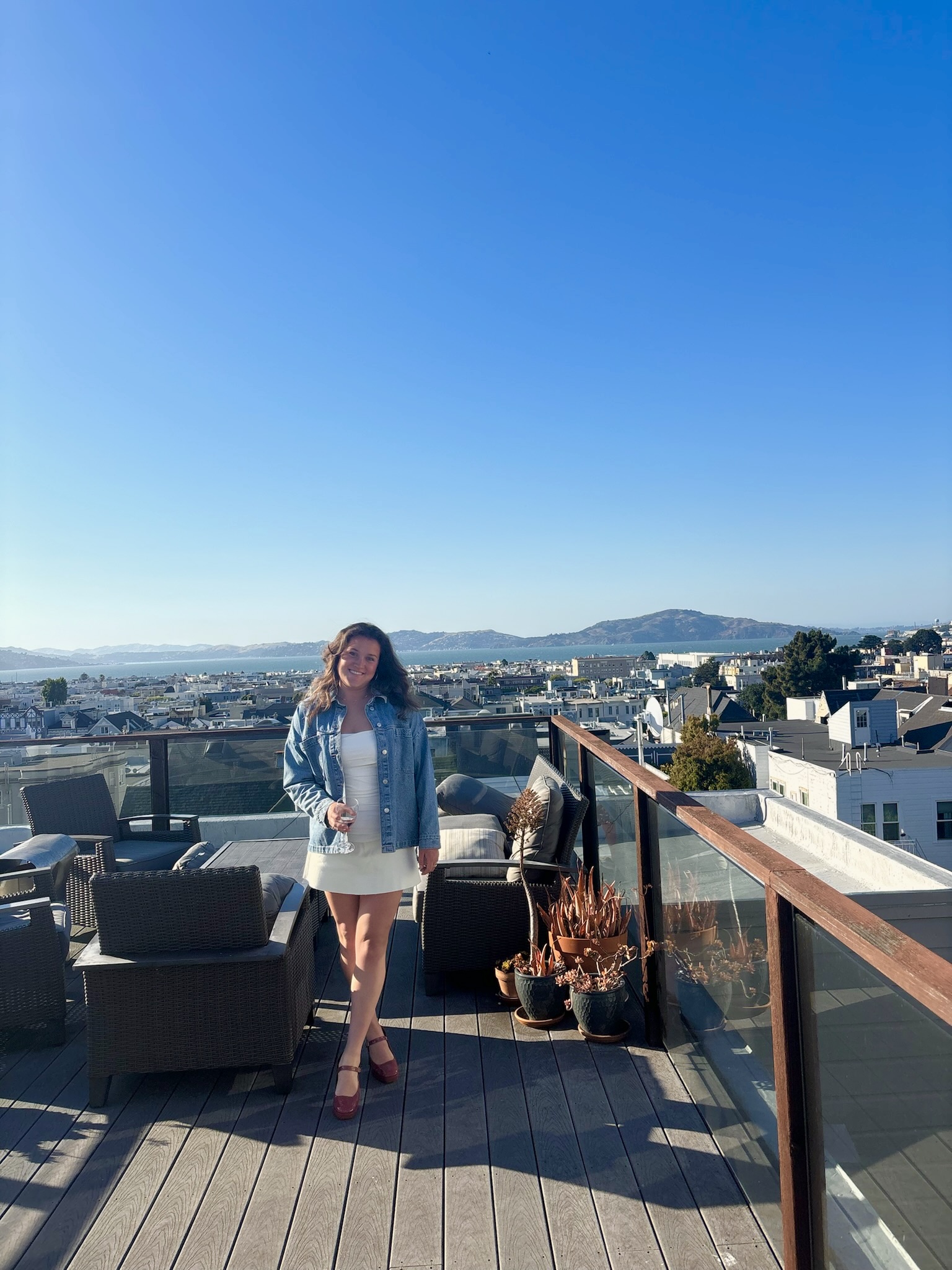 A woman in a white dress and denim jacket stands on a rooftop patio with outdoor seating, epitomizing the charm of local getaways. Her hand rests on a glass railing, and she smiles at the camera. The cityscape and distant hills unfold under a clear blue sky.