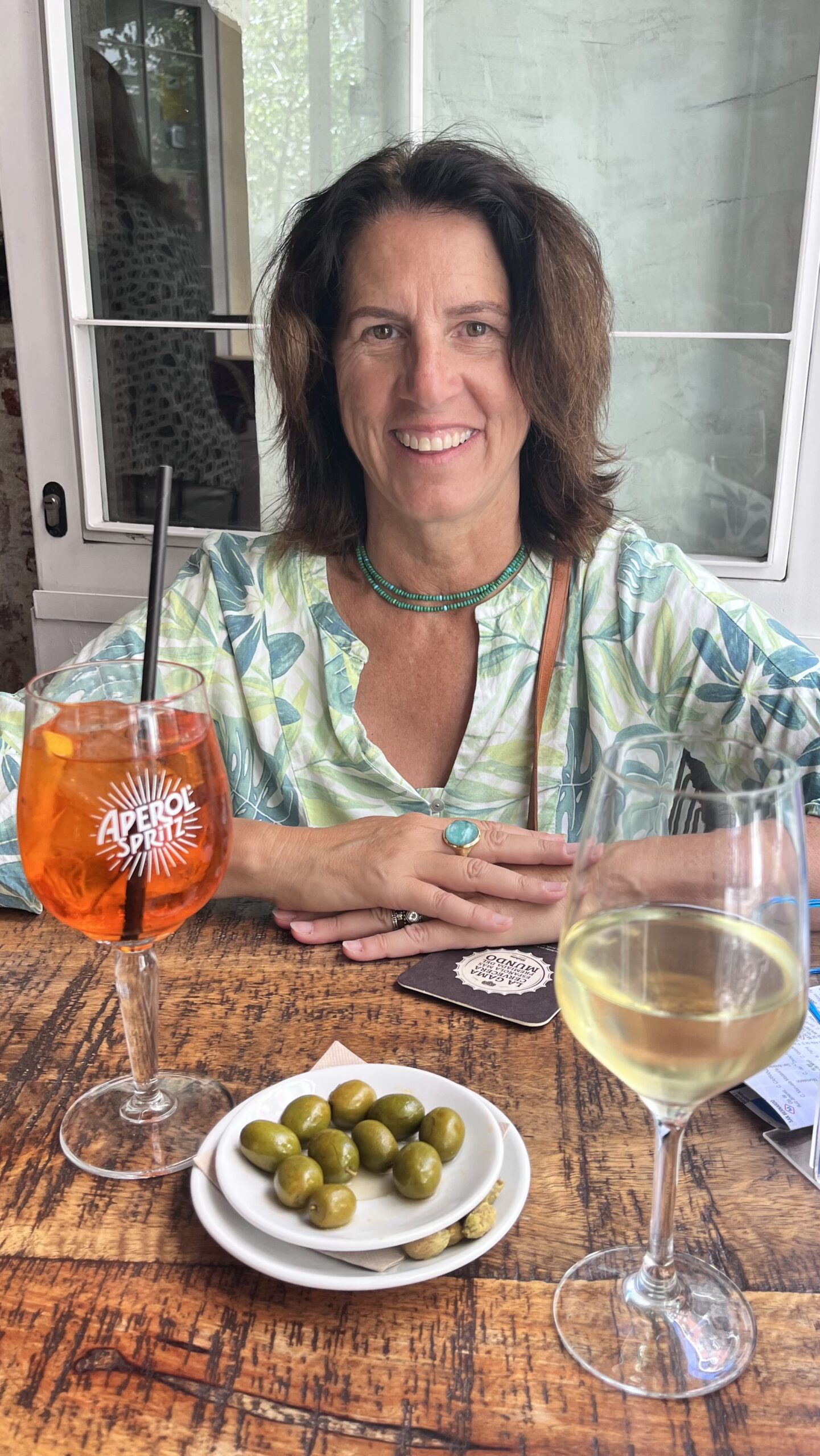 A woman with shoulder-length brown hair, wearing a patterned top, sits at a rustic wooden table adorned with green olives, an Aperol Spritz, and a glass of white wine. Smiling as she holds a drink menu, she seems like one of those local getaways food editors. A window with greenery outside frames the scene beautifully.