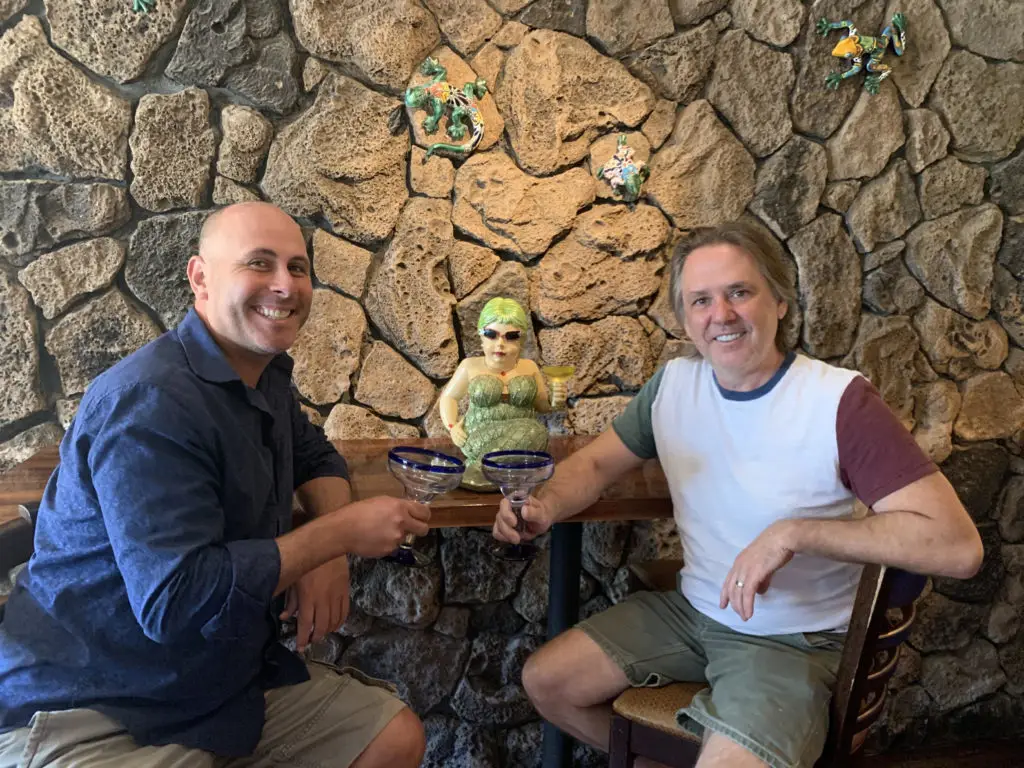 Two men sit at a table with a textured stone wall behind them, raising margarita glasses in a toast. The man on the left wears a blue shirt, while the man on the right sports a white and maroon shirt. Between them, decorations and a small statue add to the ambiance—capturing the essence of Barfly Podcast Marin.