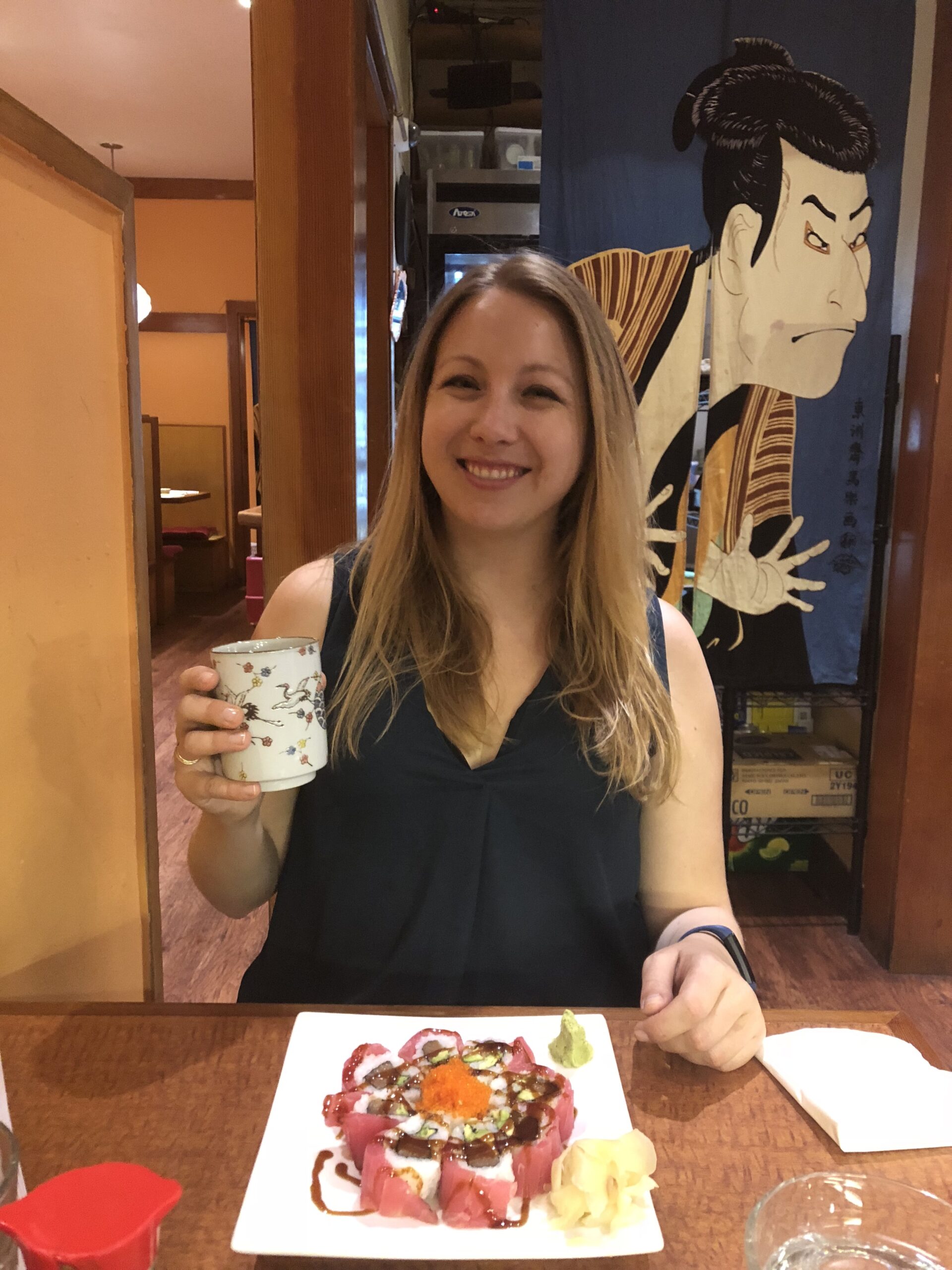 A woman with long hair sits at a table in a restaurant, smiling, and holding a cup in her left hand. In front of her is a plate of sushi. The background features Japanese-style decor and a large illustration of a traditional Japanese figure, perfect for local getaways recommended by food editors.