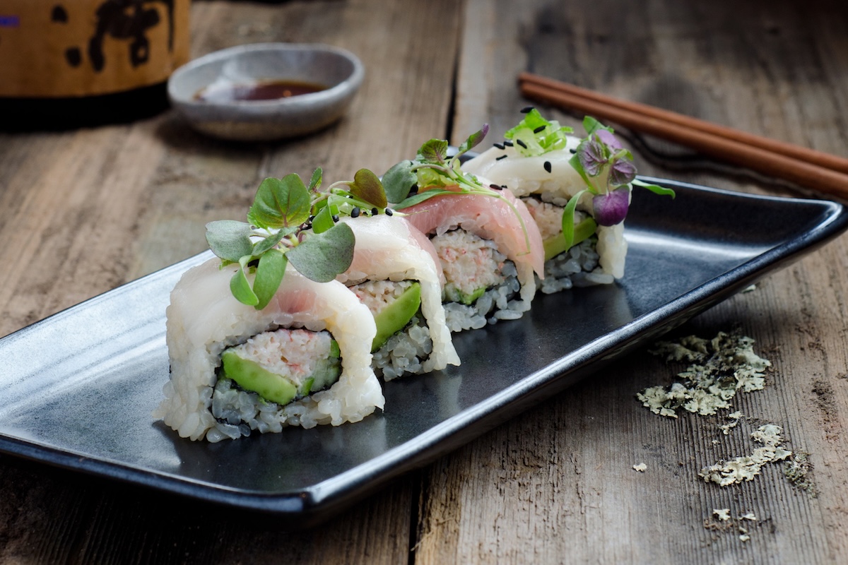 Pieces of sushi sit on a plate and are topped with microgreens for garnish. Chopsticks and a bowl of soy sauce sit next to it.