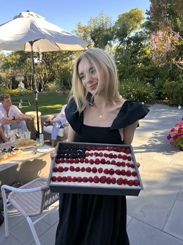 A person with long blonde hair wearing a black dress holds a tray of dessert arranged to resemble the American flag. They are standing outdoors on a patio with greenery and several people sitting under a large umbrella in the background, perfect for local getaways celebrated by food editors alike.