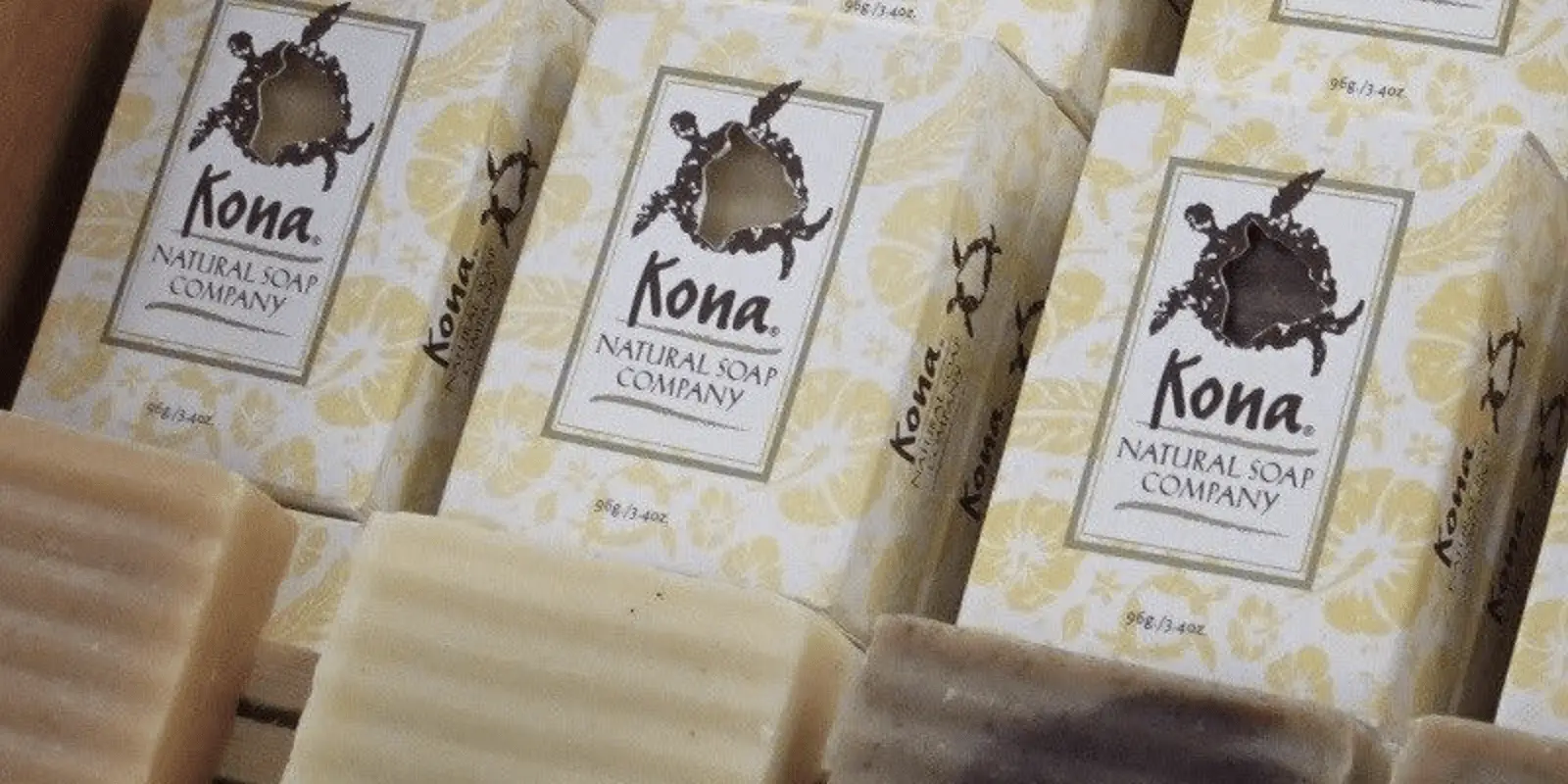 Boxes of Kona Natural Soap Company products displayed, featuring a turtle image on the packaging. Various bars of soap in front showcase different colors and textures, suggesting a variety of soap types. The background includes more Kona Natural Soap Company boxes partially visible.