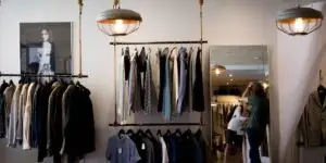 A stylishly designed clothing store with hanging lights. Racks display various shirts, jackets, and pants. A large mirror reflects a person browsing clothing in the background. A framed photo of a model hangs on the wall to the left.