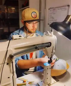 A person wearing glasses and a beige Tommy Breeze cap with a mountain logo sews apparel using a sewing machine. The workspace is illuminated by a lamp, and there are tools and a whiteboard in the background. The individual is focused on their task.