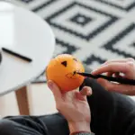 A person with glasses decorates an orange with a black marker, drawing a face with triangular eyes and a smiling mouth. Seated next to a white table adorned with more oranges and markers, they showcase creative pumpkin carving alternatives. A patterned rug is visible in the background.