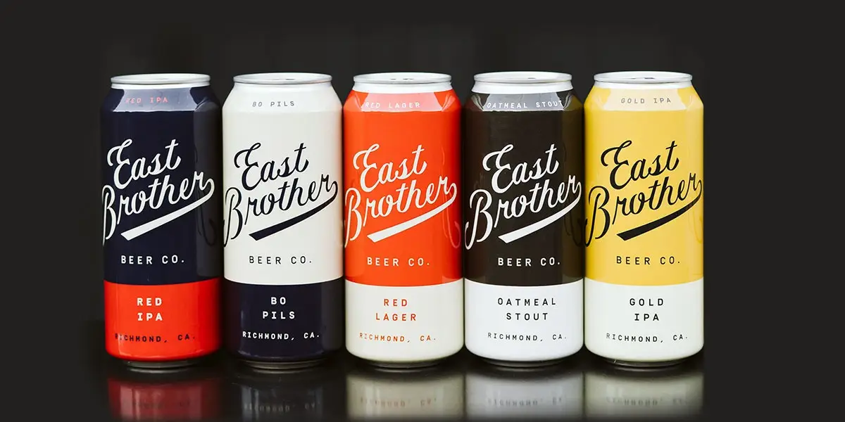 Five cans of East Brother Beer are lined up in a row against a black background. The cans, from left to right, are labeled as Red IPA, Bo Pils, Red Lager, Oatmeal Stout, and Gold IPA, showcasing the diverse flavors and distinct designs that East Brother Beer is known for.