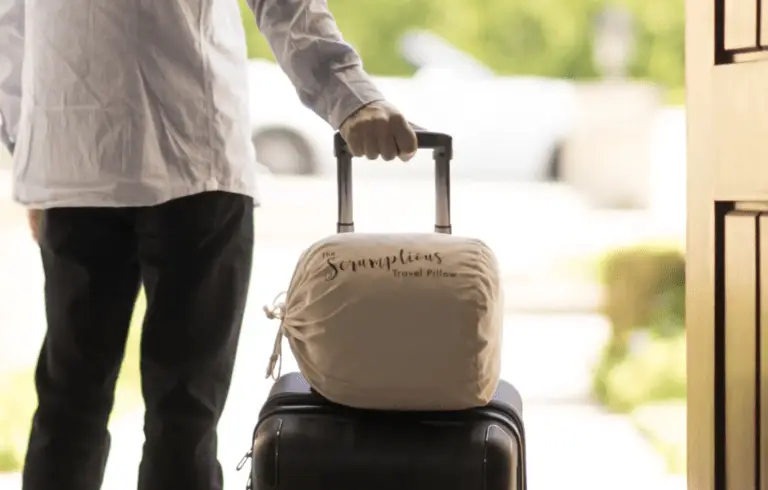 A person wearing a white shirt is seen from the waist down, holding the handle of a suitcase. On top of the suitcase are travel sleep essentials, including a travel pillow with the brand name "Scrumptious." The scene appears to be at an entrance or exit with an outdoor background blurred.