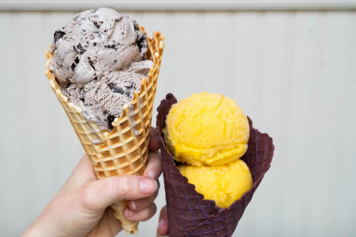 Two people hold ice cream cones, one a plain waffle cone with cookies and cream ice cream and the other mango sorbet in a chocolate waffle cone.