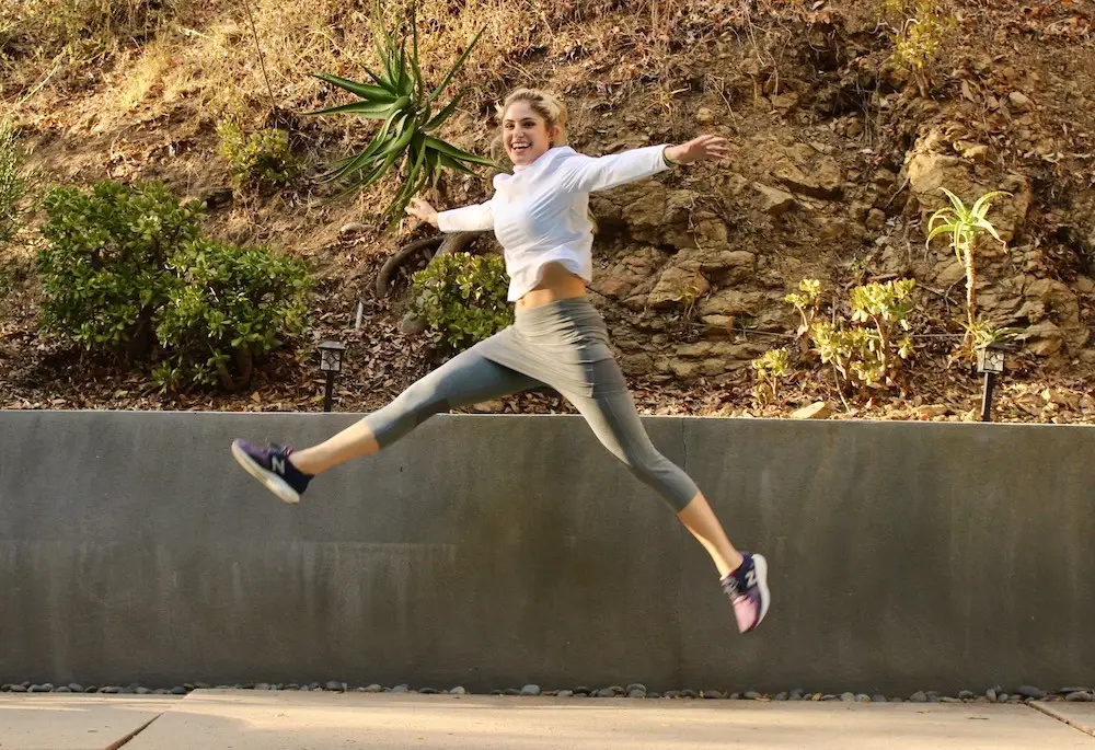 A person wearing a white top, gray leggings, and black sneakers from Sol Sister Sport is captured mid-air while leaping outdoors. They have their arms stretched out wide and appear to be joyful. The background includes a concrete wall, greenery, and a rocky hillside.