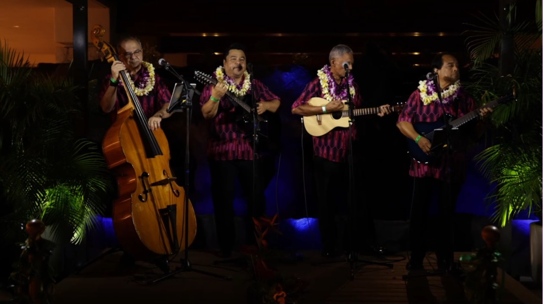 Four musicians wearing leis and matching patterned shirts perform on stage at the Outrigger music venue. The group consists of a double bass player, a singer holding a ukulele, a guitar player, and another string instrument player. The dimly lit stage features tropical plants against a dark background, evoking a Hawaii home feel.