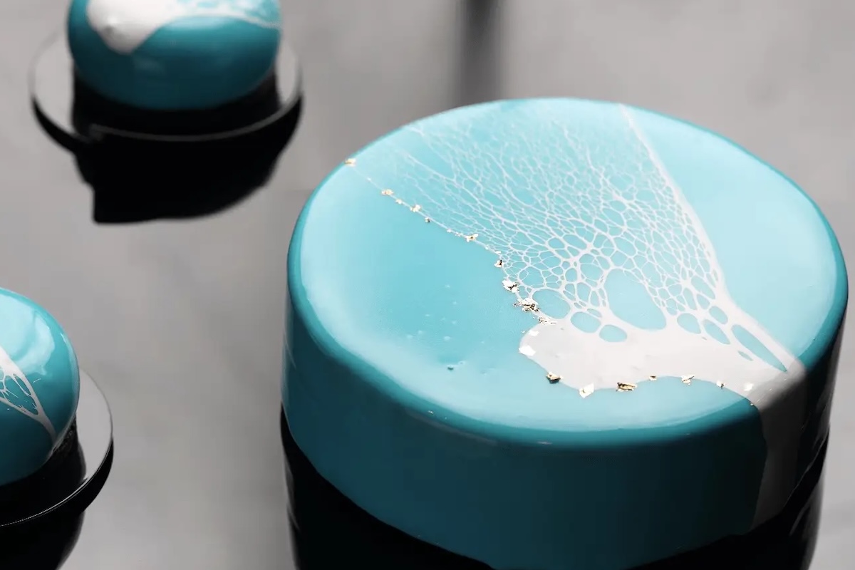 A large round blue cake with a white, lacy design on top sits on a reflective black surface next to a smaller, similarly decorated round blue dessert. The cakes appear glossy, with a smooth texture and delicate gold leaf accents— truly among the best desserts in the Bay Area.