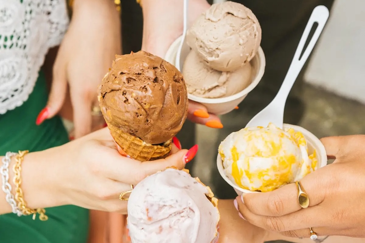 Four hands holding various ice cream treats, perhaps some of the Best Desserts in the Bay Area. The treats include a chocolate ice cream cone, a scoop of beige ice cream in a bowl with a spoon, a yellow and white swirled ice cream in a cup, and a pinkish ice cream cone. The background is out of focus.