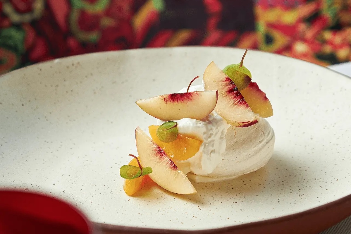 A contender for the Best Desserts in the Bay Area, this gourmet treat features a scoop of vanilla ice cream or sorbet elegantly topped with thin, artfully arranged slices of peach and small green garnish leaves, all served on a minimalist speckled ceramic plate against vibrant, colorful patterns.