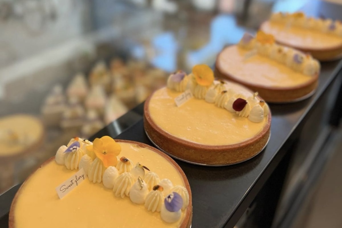 A row of four lemon tarts is displayed on a bakery counter. Each tart, topped with cream dollops and delicate edible flowers, epitomizes the culinary craftsmanship found in top "Where to Eat Bay Area" spots. In the background, more baked goods are partially visible out of focus.