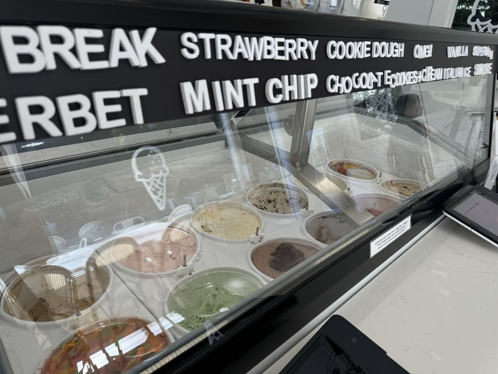 A display case at an ice cream shop filled with various flavors of ice cream and sorbet, showcasing some of the Best Desserts in the Bay Area. Visible flavors include Break, Strawberry, Cookie Dough, Mint Chip, Chocoat, and more. The ice cream tubs are arranged in a neat row with an illustrated cones visible.