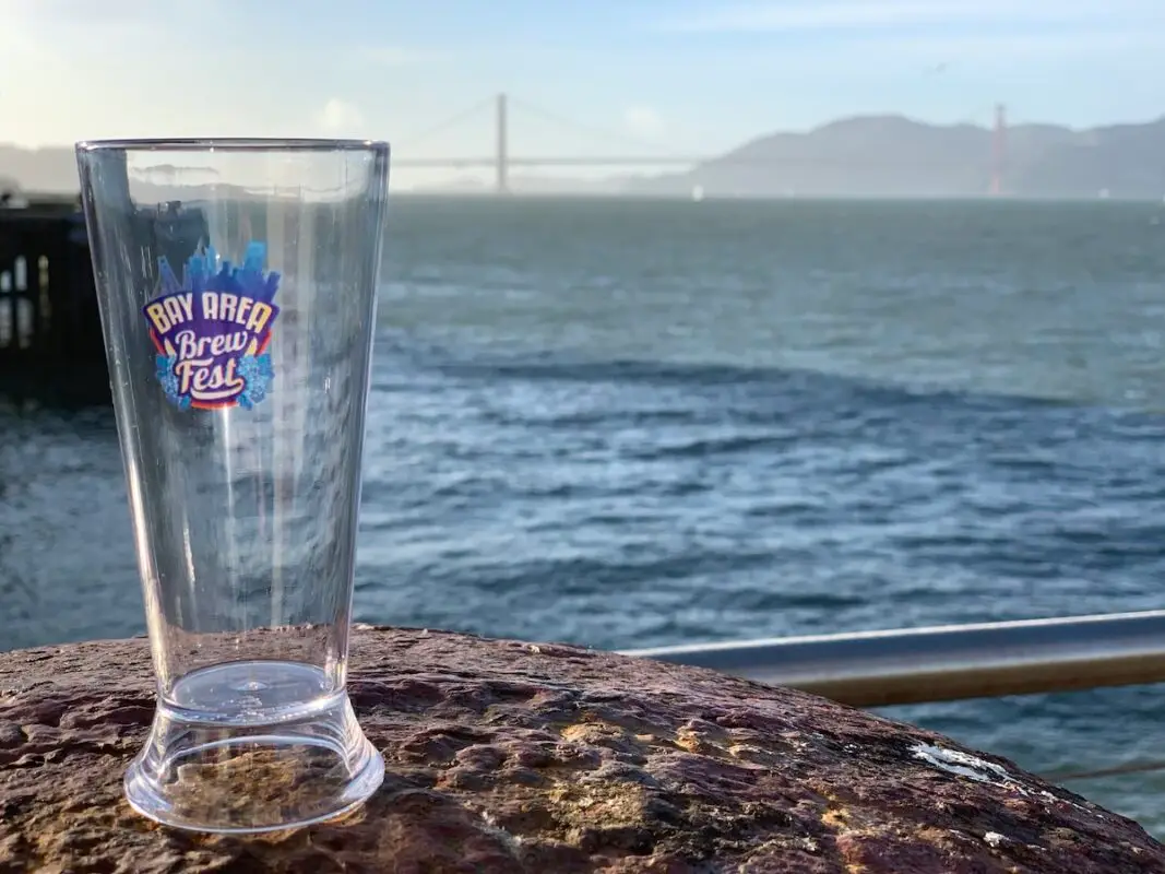 Empty beer pint glass on fence above the bay and Golden Gate Bridge in distance, labeled "Bay Area Brew Fest" for the festival in San Francisco, California