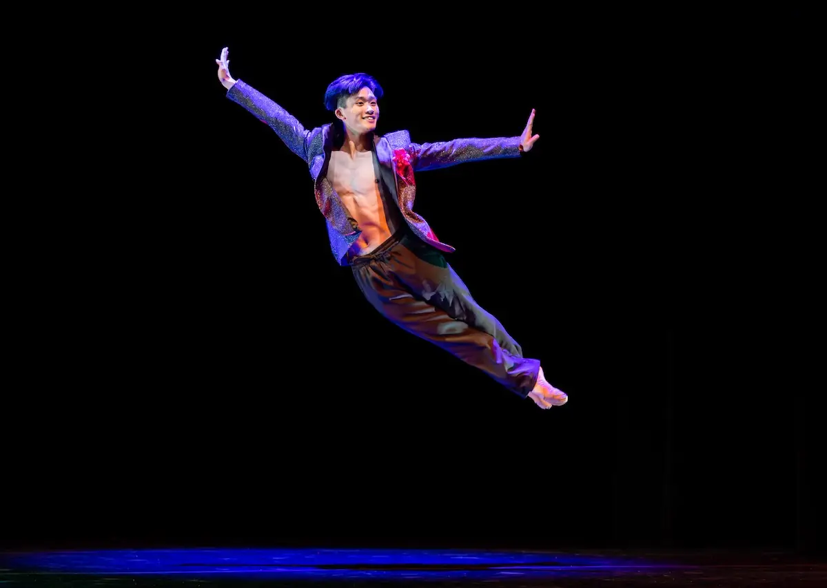 A male dancer dressed in a sparkly jacket and dark pants performs a mid-air leap against a black background. His left arm and right leg are extended elegantly, and he smiles while looking slightly upward. The stage floor has a subtle blue glow, capturing one of the best things to do in San Francisco in April.