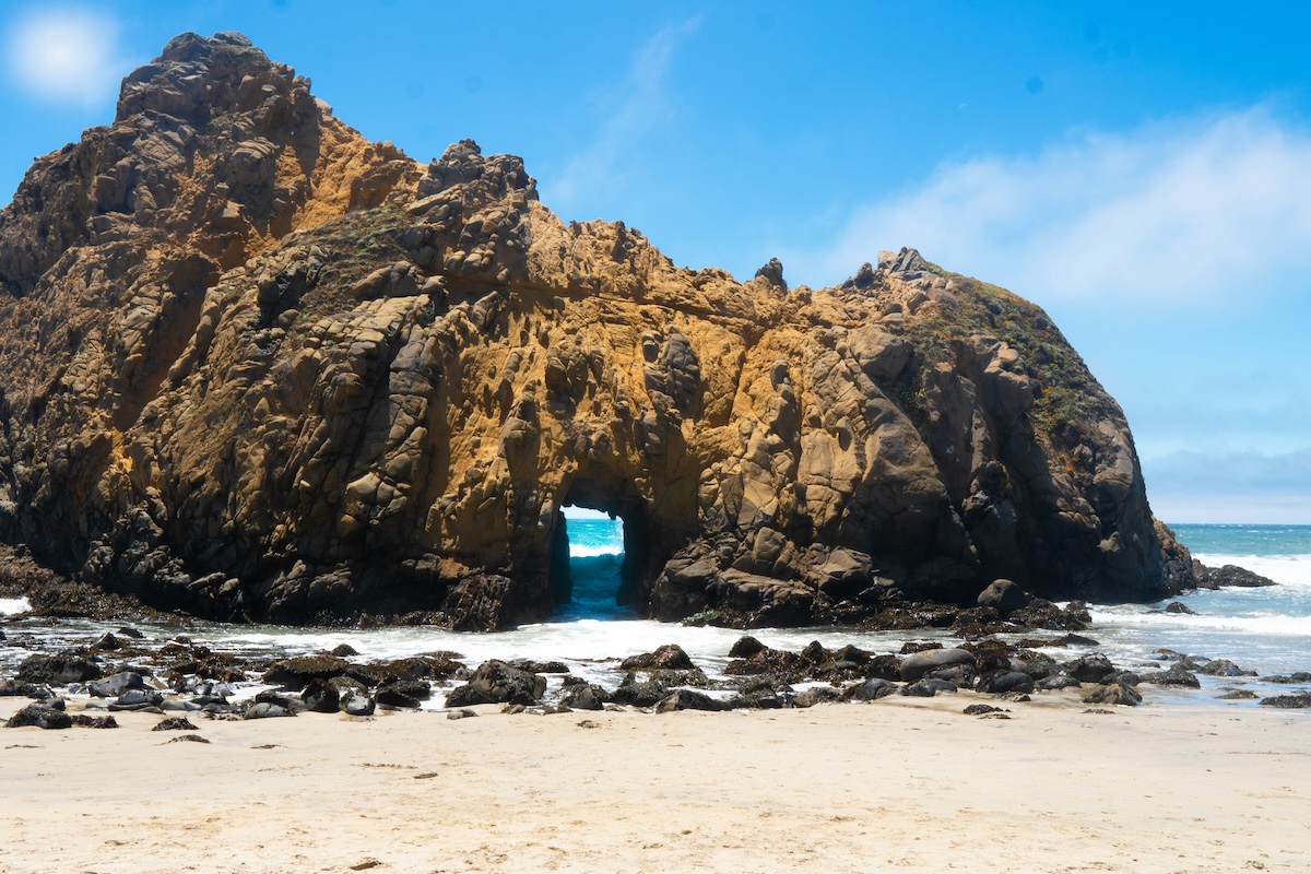 A giant rock arch sits on the edge of water on a beach against a blue sky.