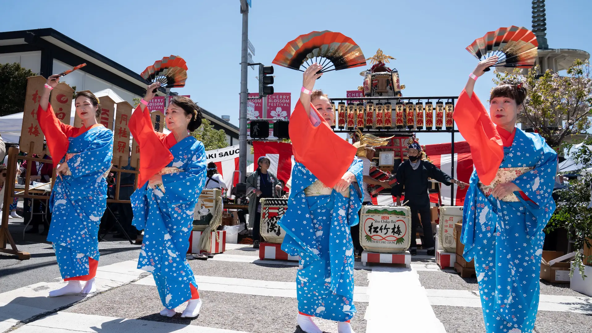 Four women dressed in vibrant blue and red traditional Japanese attire perform a fan dance outdoors in one of the best things to do in San Francisco this April. They are in sync, fans raised, before an ornate structure with musicians playing instruments against a clear blue sky background. The scene is festive and lively.