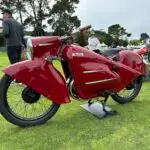 A vintage red motorcycle with a streamlined, Art Deco-inspired design is displayed on a manicured lawn. The bike features the "Miller" logo on its side. Several people in the background are admiring various motorcycles at what appears to be an outdoor event, one of the best things to do on the Monterey Peninsula May 2024.